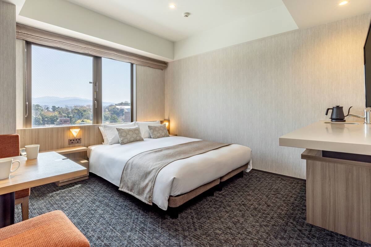 A hotel room with a large bed, a desk with a chair, a window with a view, and a bathroom area.