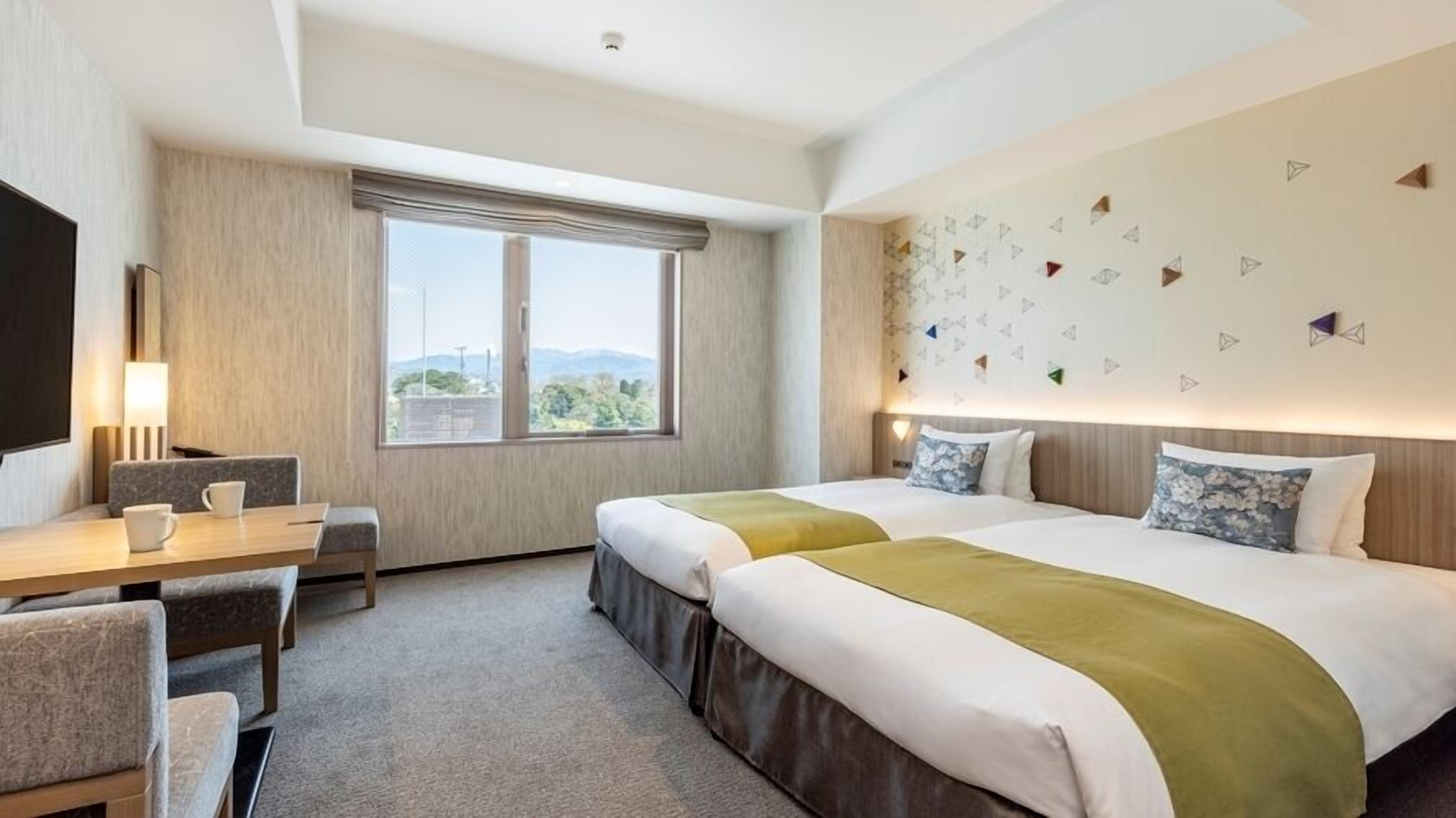 A hotel room with two beds, a desk, a chair, and a large window.