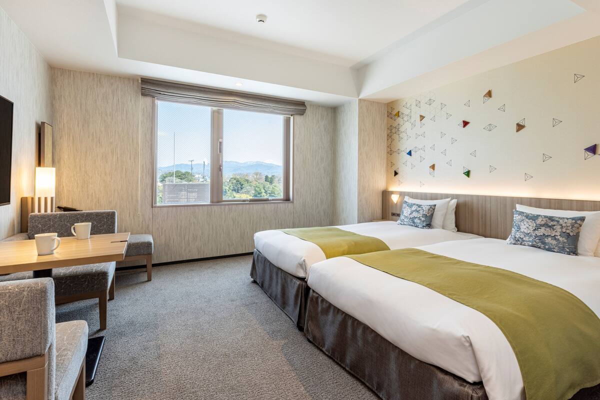 A hotel room with two beds, a desk, a chair, a large window with a view, and a wall with geometric patterns.