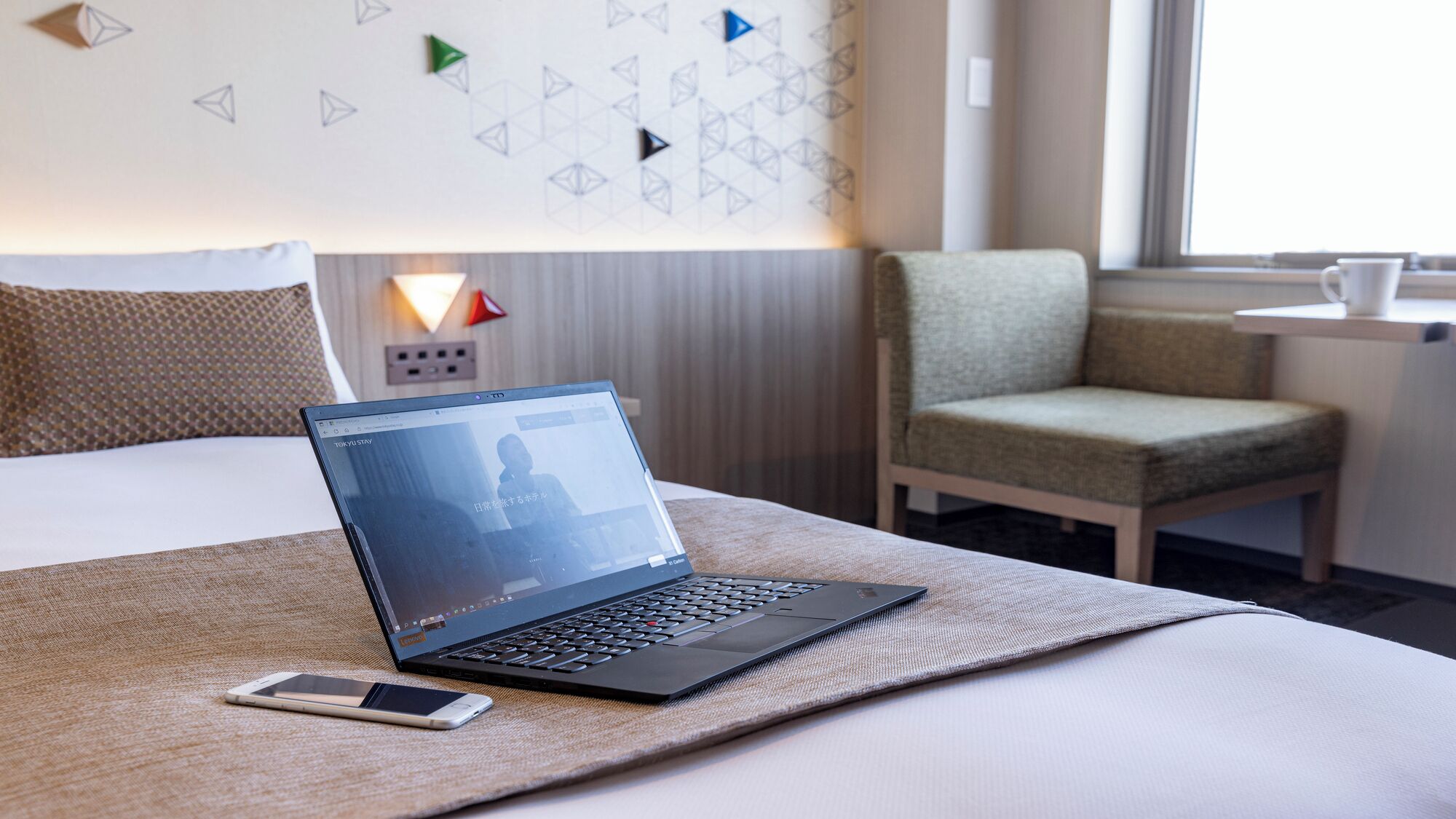 A laptop on a bed with a smartphone beside it, a chair, and a wall with geometric patterns.