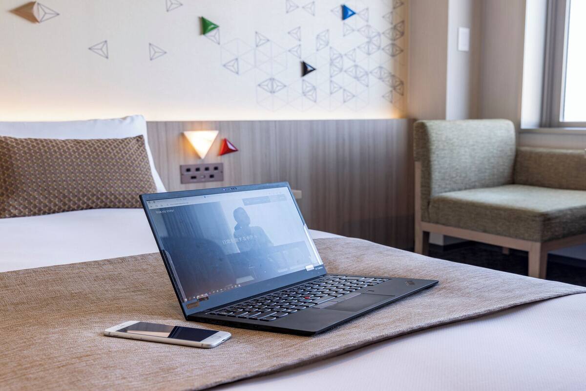 A laptop on a bed with a smartphone beside it, a chair, and a wall with geometric patterns.