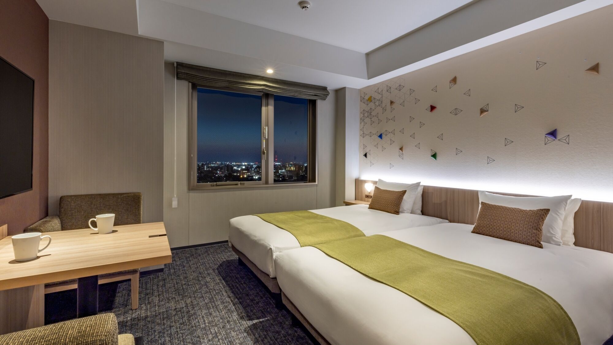 A hotel room with a large bed, a desk with a chair, a window with a view of the city at night, and a wall with geometric patterns.