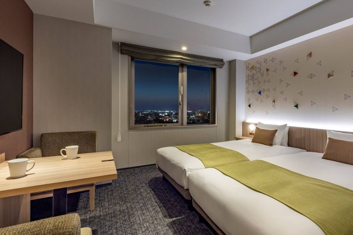 A hotel room with a large bed, a desk with a chair, a window with a view of the city at night, and a wall with geometric patterns.
