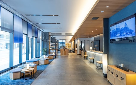 A modern hotel lobby with a long reception desk, seating area, and large windows offering views of the street.