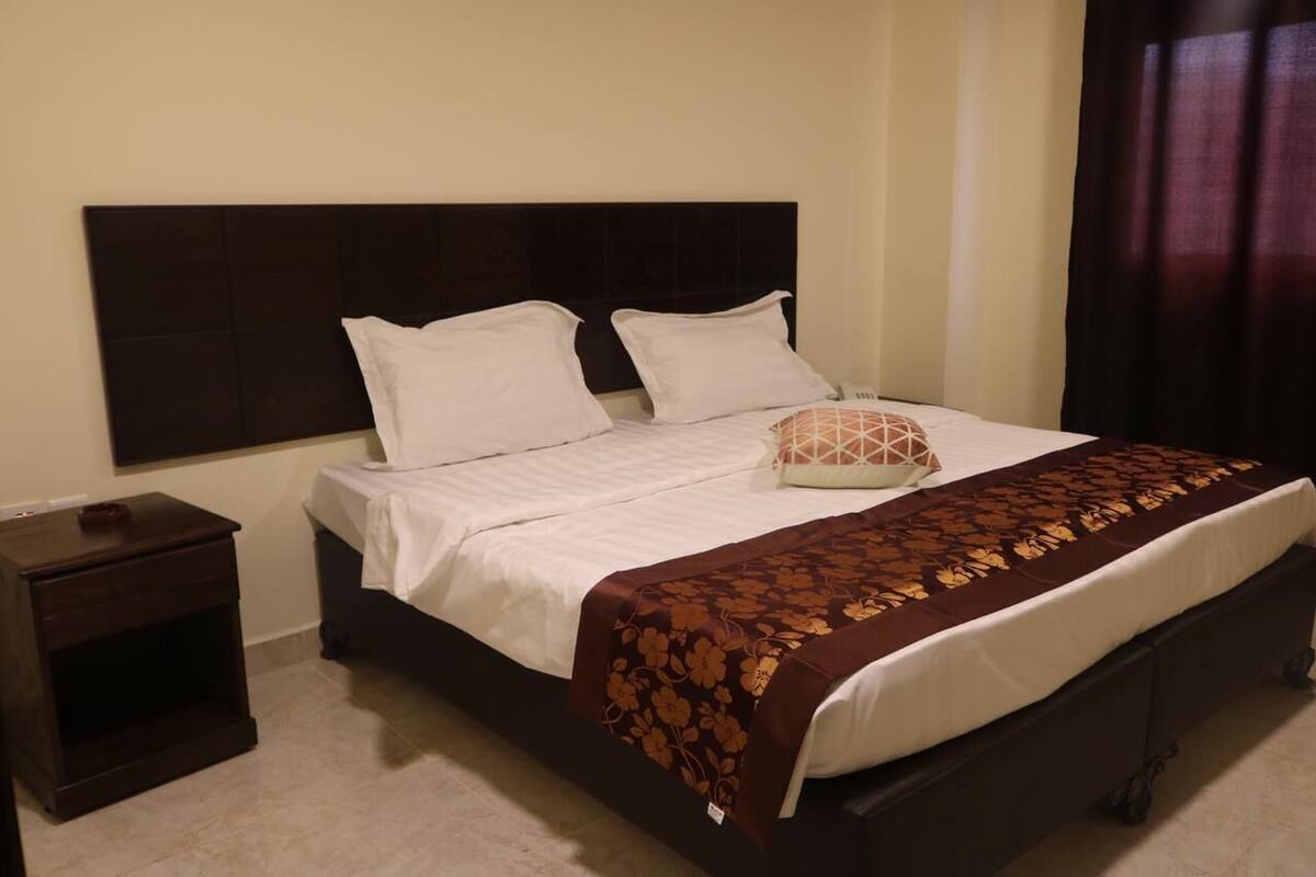 A neatly made bed with white linens and a brown floral bedspread, a dark headboard, and a small wooden nightstand with a drawer.