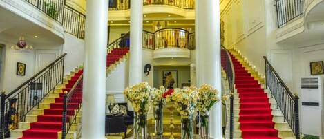 A grand lobby with a central round table, two red-carpeted staircases, and a chandelier.