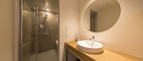 Un baño moderno con cabina de ducha de vidrio, tocador de madera con lavamanos y espejo redondo.