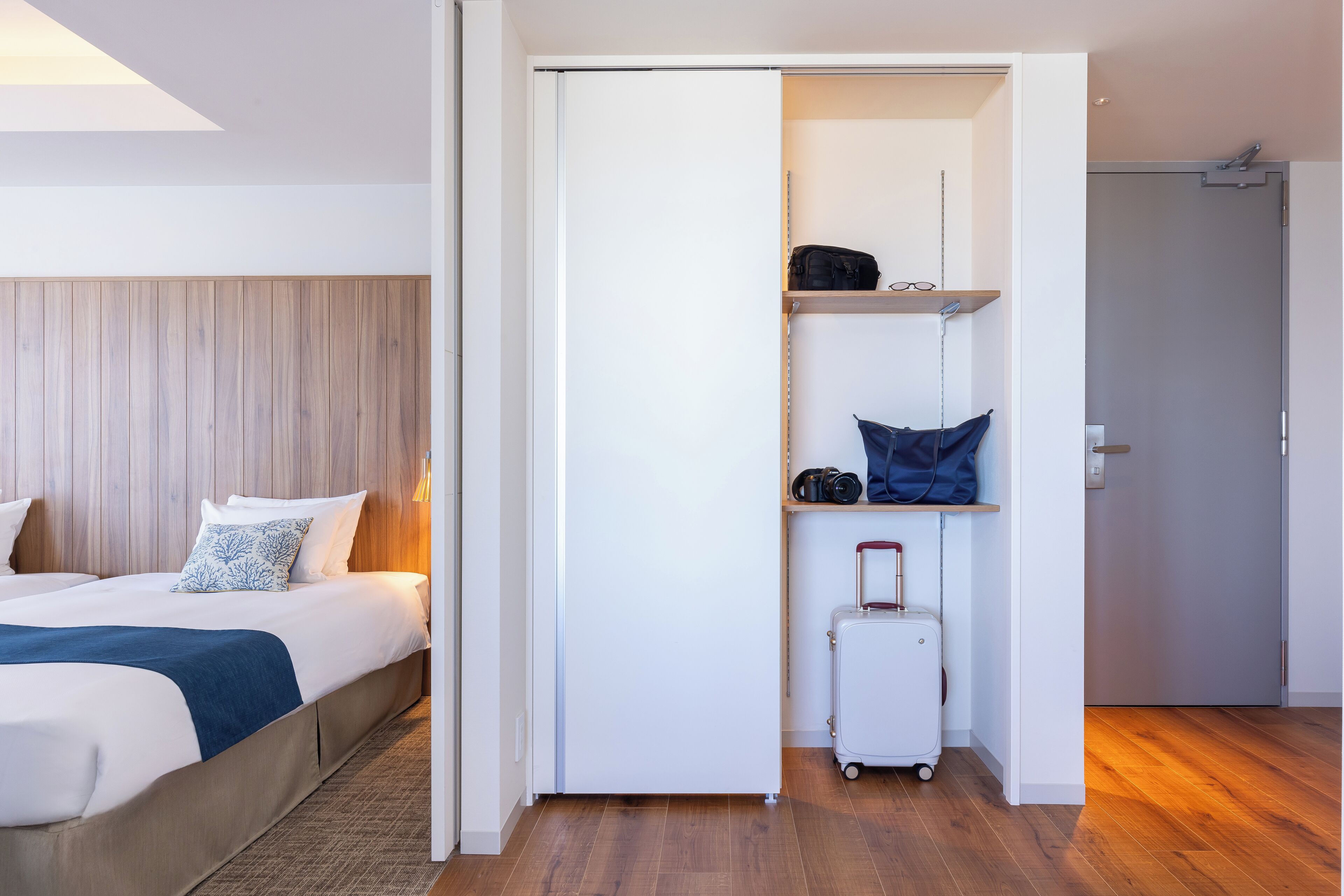 A hotel room with a bed, a wardrobe, a suitcase, and a shelf with personal items.