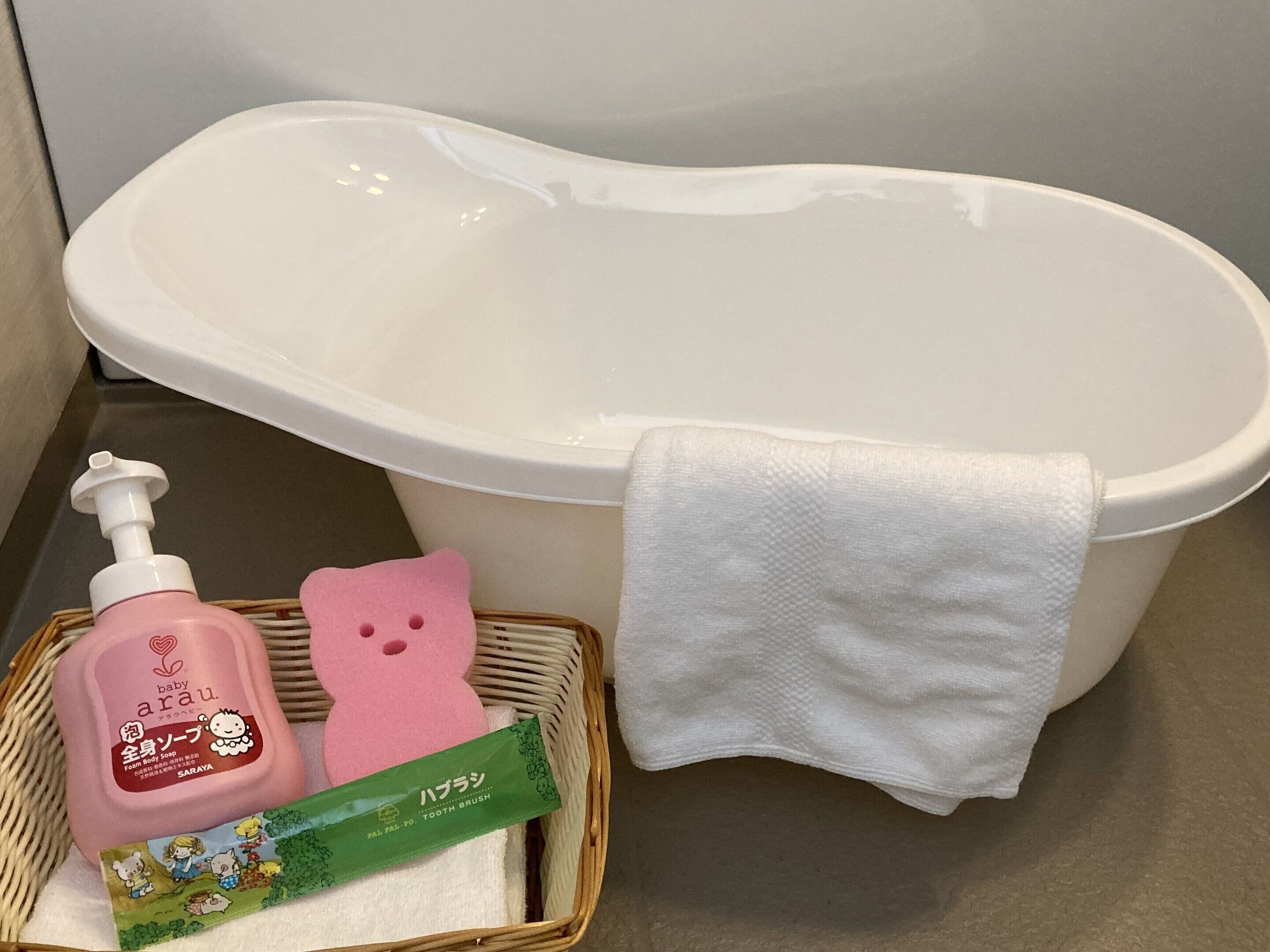 A bathroom with a white bathtub, a white towel, a pink baby soap dispenser, a pink baby bath sponge, and a green toothbrush package.