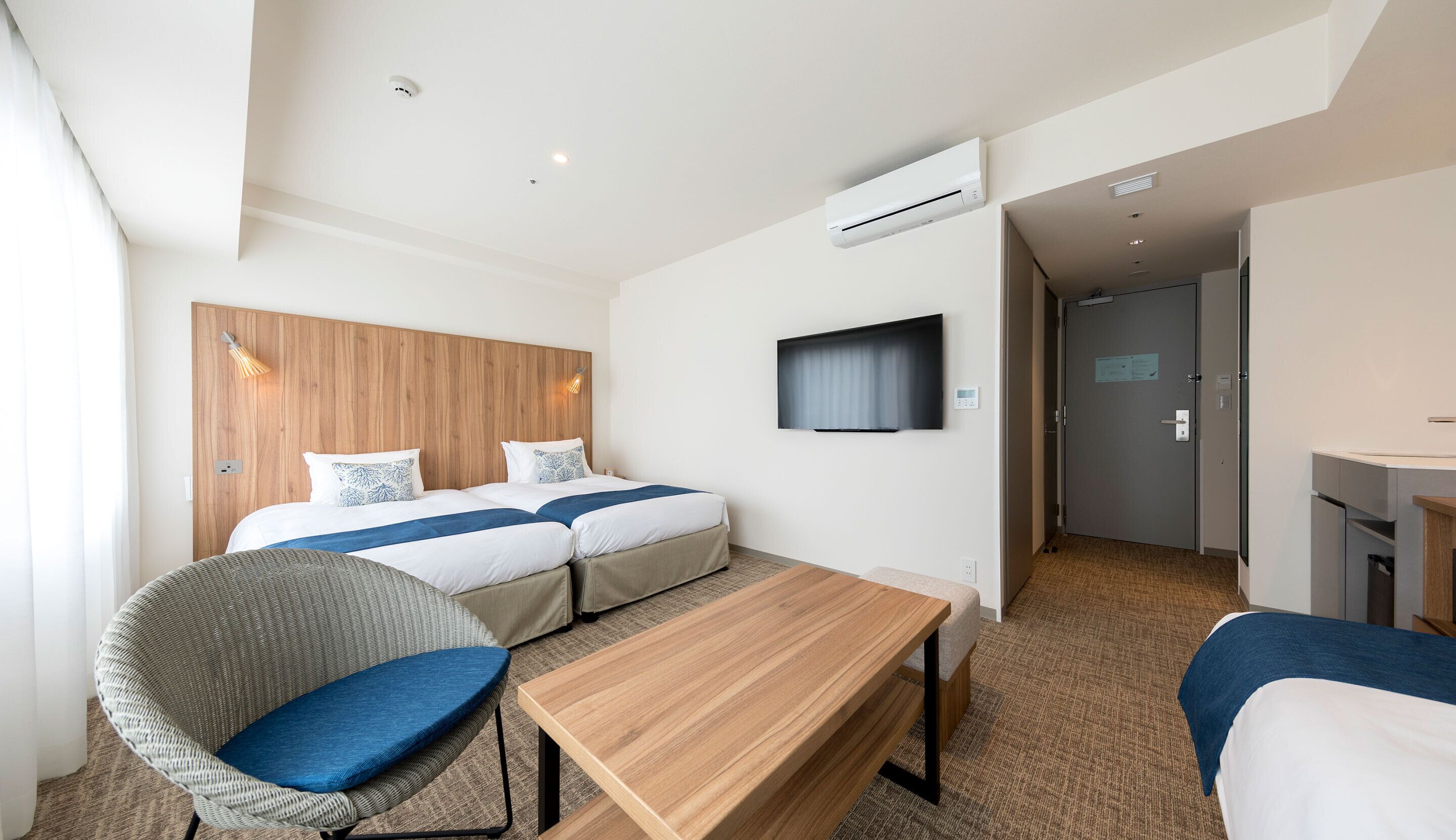 Habitación de hotel con dos camas, televisión, aire acondicionado, cabecero de madera y zona de estar con mesa y sillas.