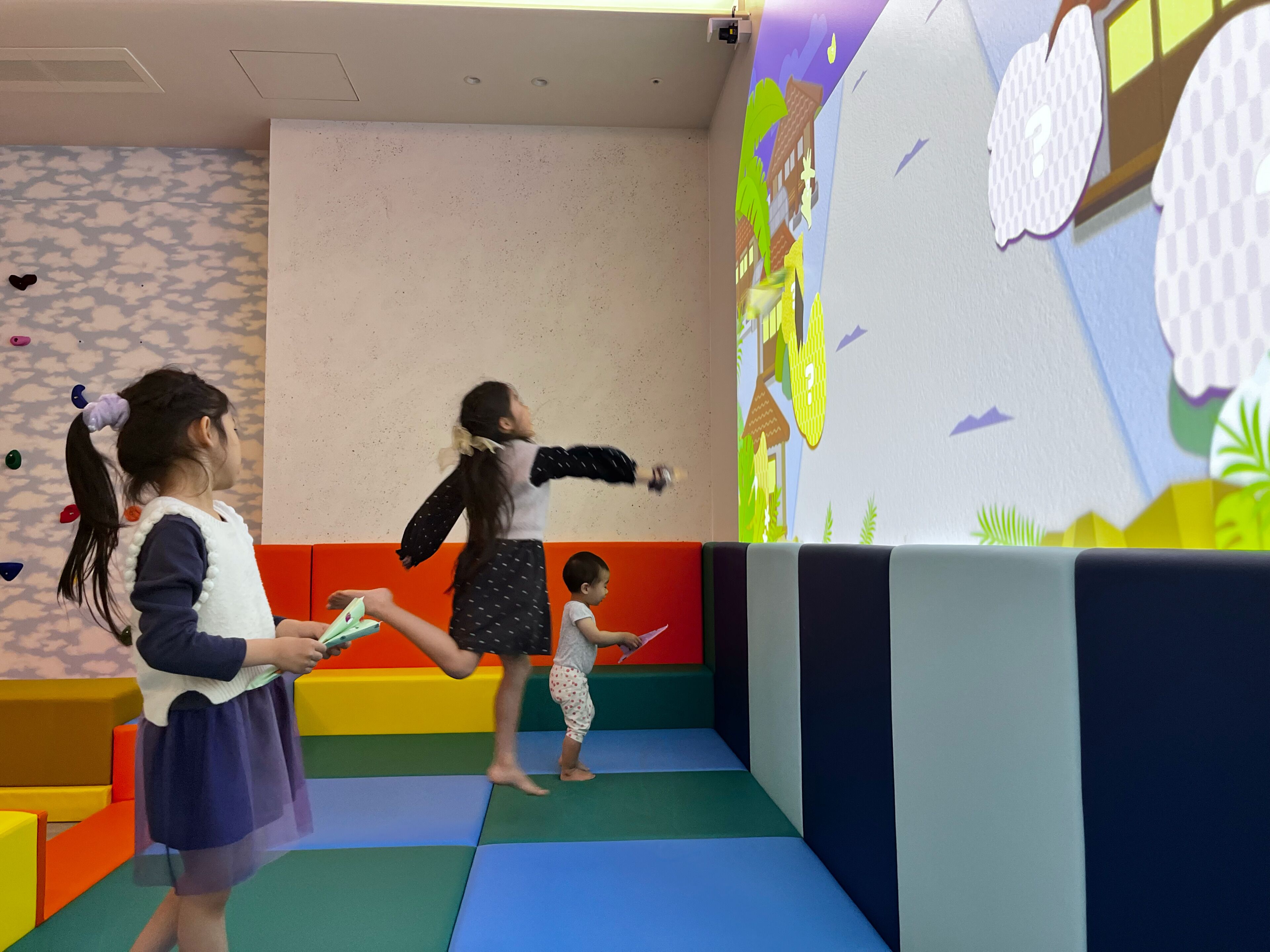 Bambini che giocano in un'area giochi al coperto colorata, con strutture in gomma morbida e una parete decorata con un paesaggio naturale.
