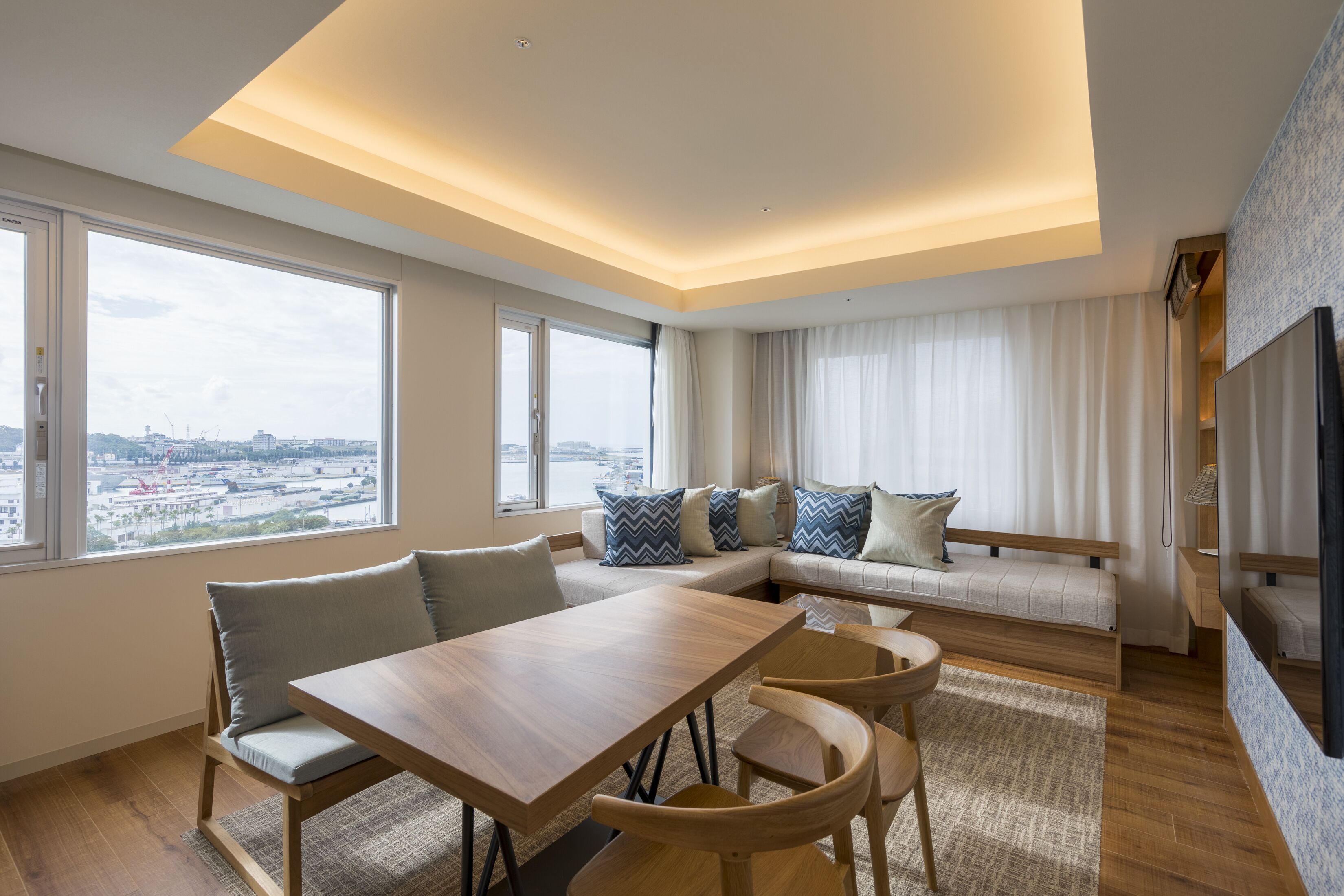 A modern living room with a wooden dining table, cushioned seating, a sofa with patterned pillows, and large windows offering a city view.