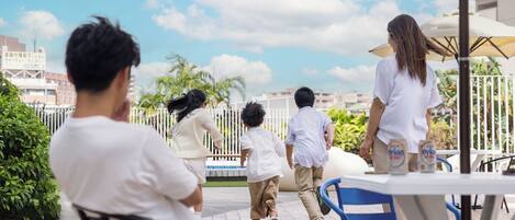 Um grupo de pessoas caminhando em um terraço com vista para a cidade, mesas ao ar livre e uma grade.