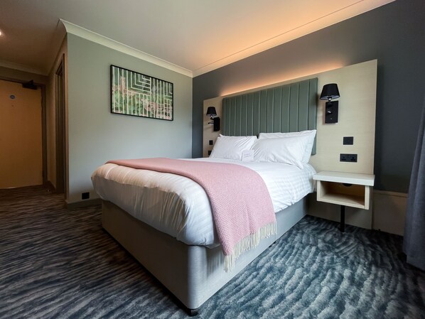 A neatly made bed with a pink throw, a headboard with built-in lighting, and a wall-mounted artwork.
