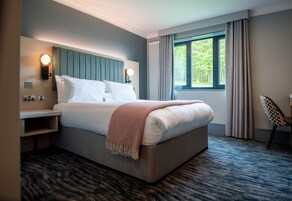 Una habitación de hotel moderna con una cama grande, una mesita de noche y una silla, además de un ventanal con cortinas.