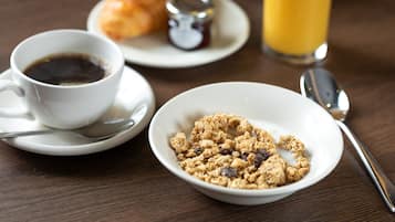 A breakfast setting with a cup of coffee, a bowl of granola, a croissant, a glass of orange juice, and a spoon on a wooden table.