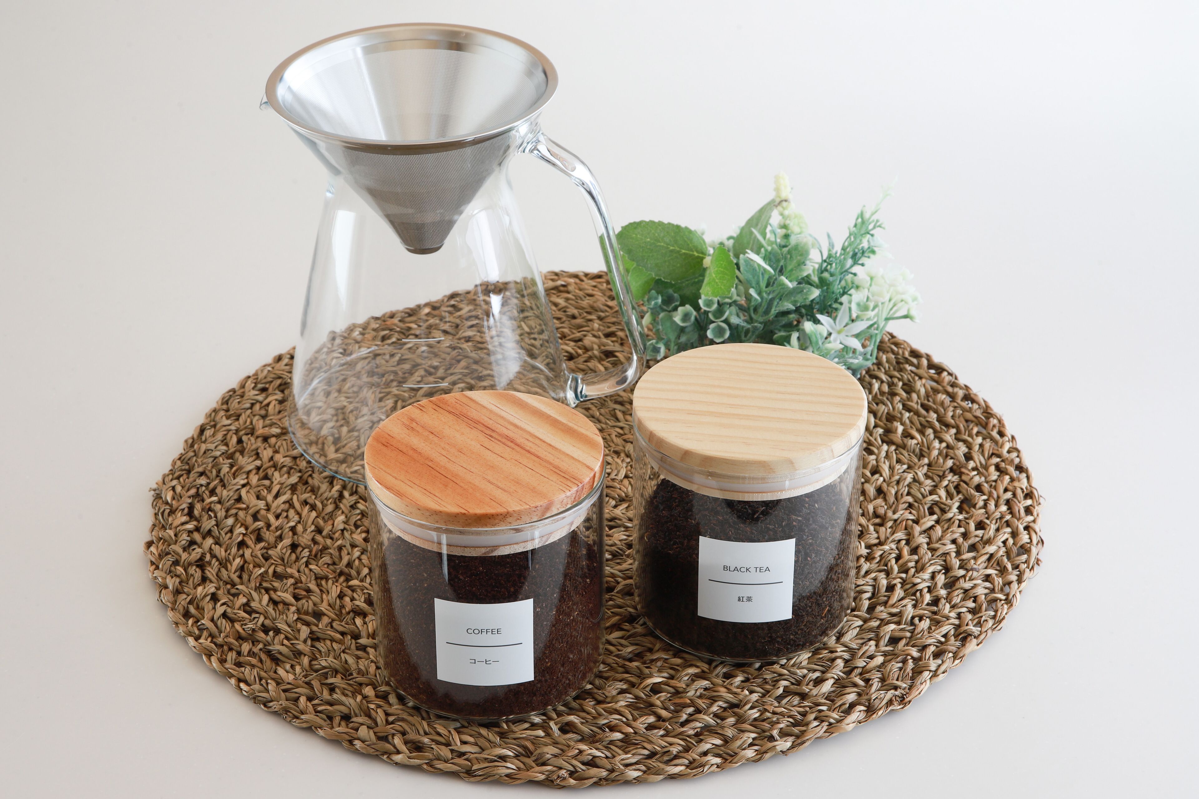 Two glass jars with wooden lids, one labeled "COFFEE" and the other "BLACK TEA," placed on a woven mat.