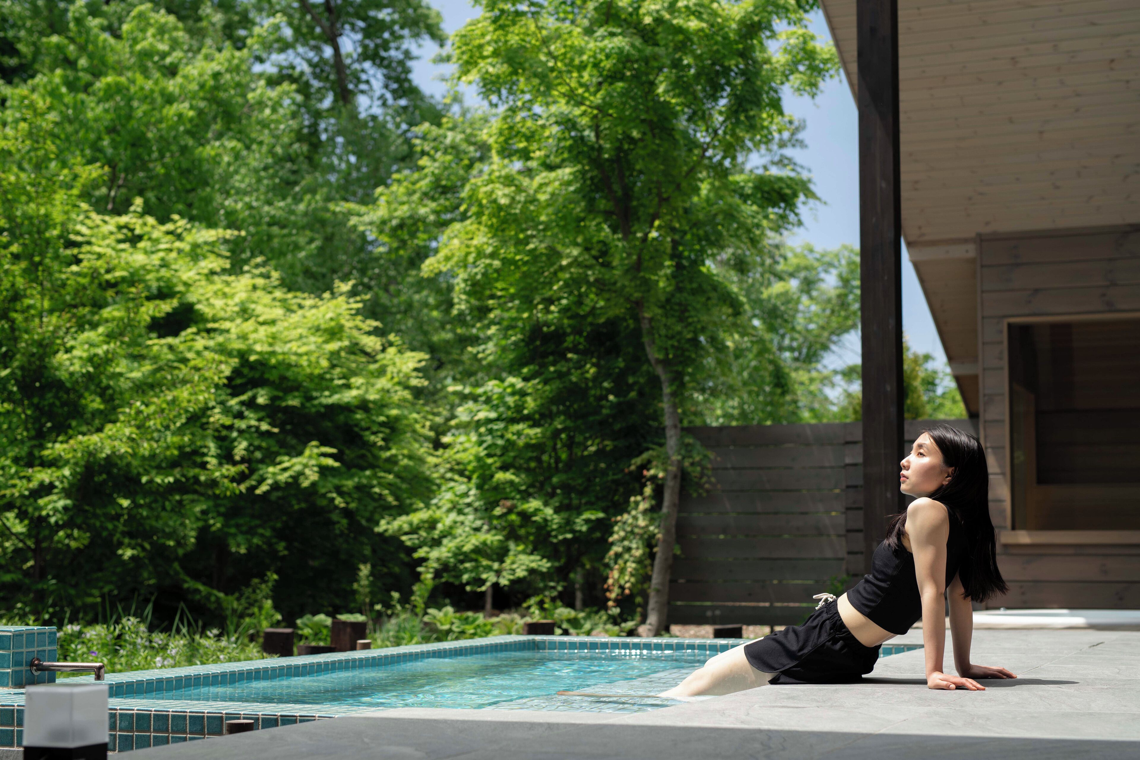 Une personne effectue une posture de yoga au bord d’une piscine, sur une terrasse en bois, avec une végétation luxuriante en arrière-plan.