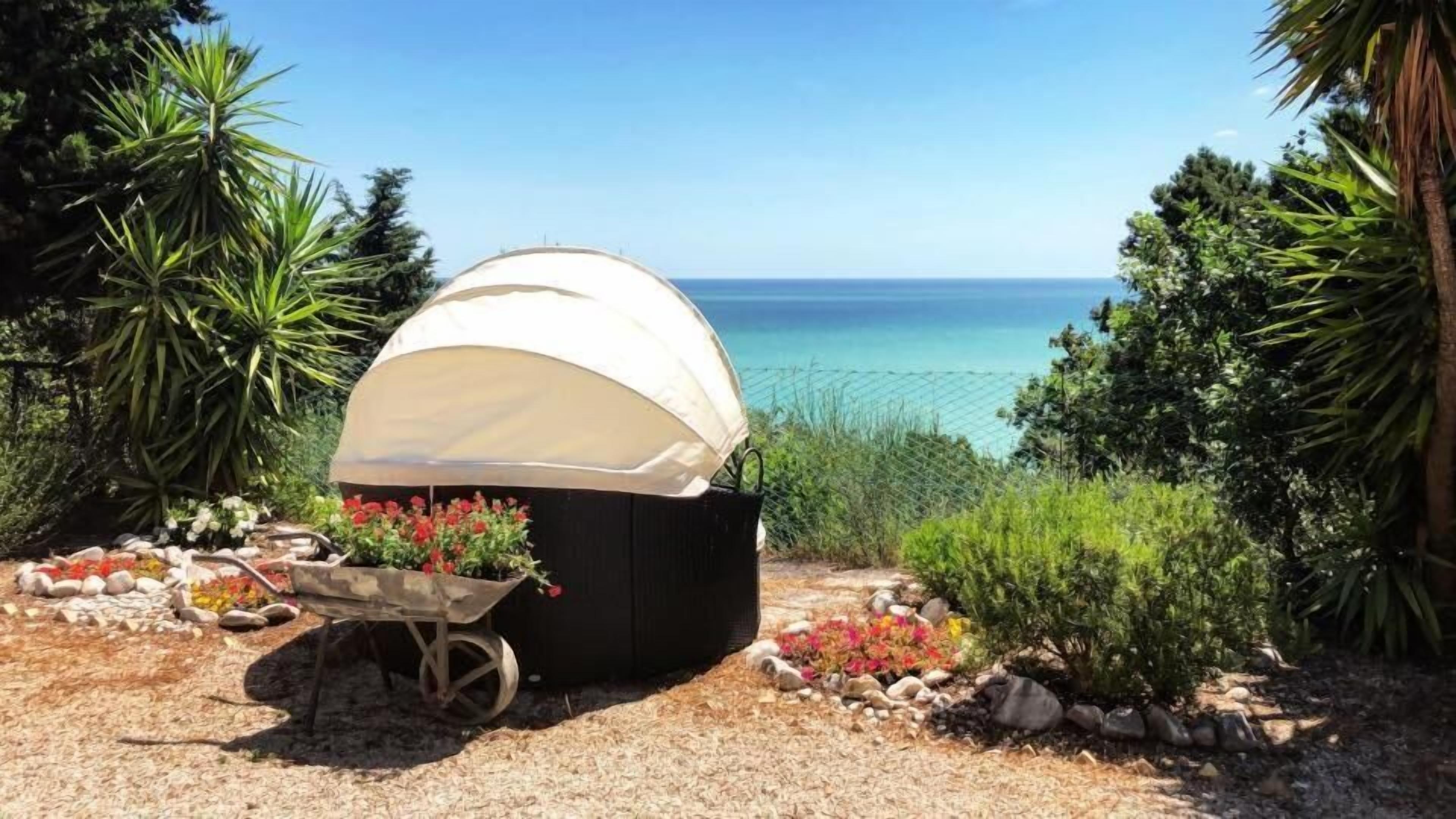 A covered outdoor seating area with a view of the sea, surrounded by plants and rocks.