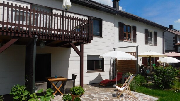 A white house with a wooden balcony, stone patio, and outdoor seating area with umbrellas. - Ferienapartments Ida (Lorch)