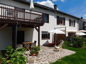 A white house with a wooden balcony, stone patio, and outdoor seating area with umbrellas.