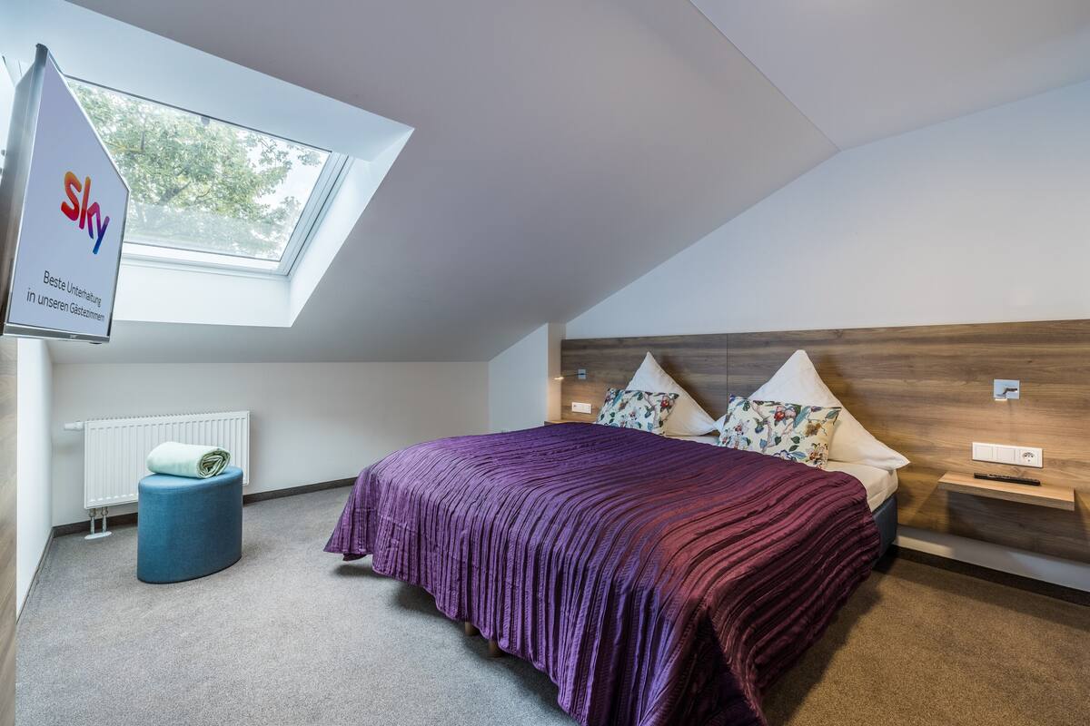 Un dormitorio con tragaluces, una cama con colcha púrpura, un reposapiés azul, un radiador blanco y un cabecero de madera.