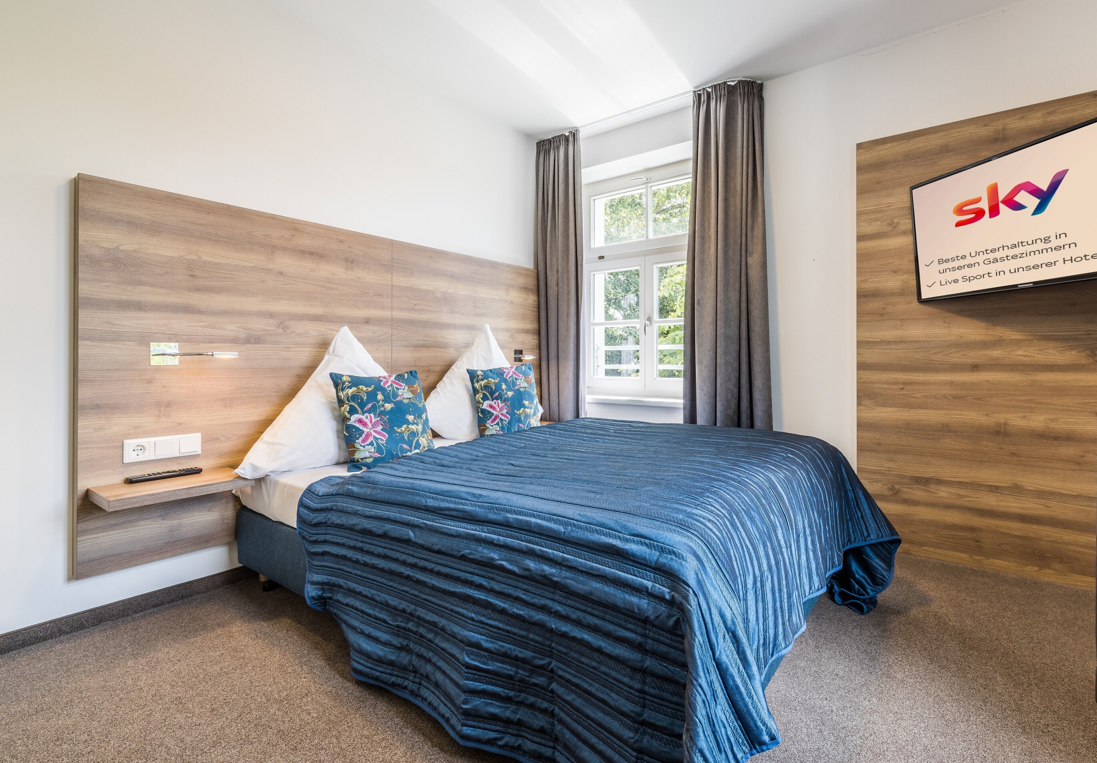 Ein Hotelzimmer mit einem Holzkopfteil, einem großen Bett mit blauer Bettdecke, einem Nachttisch mit Fernbedienung und einem Fenster mit Vorhängen.