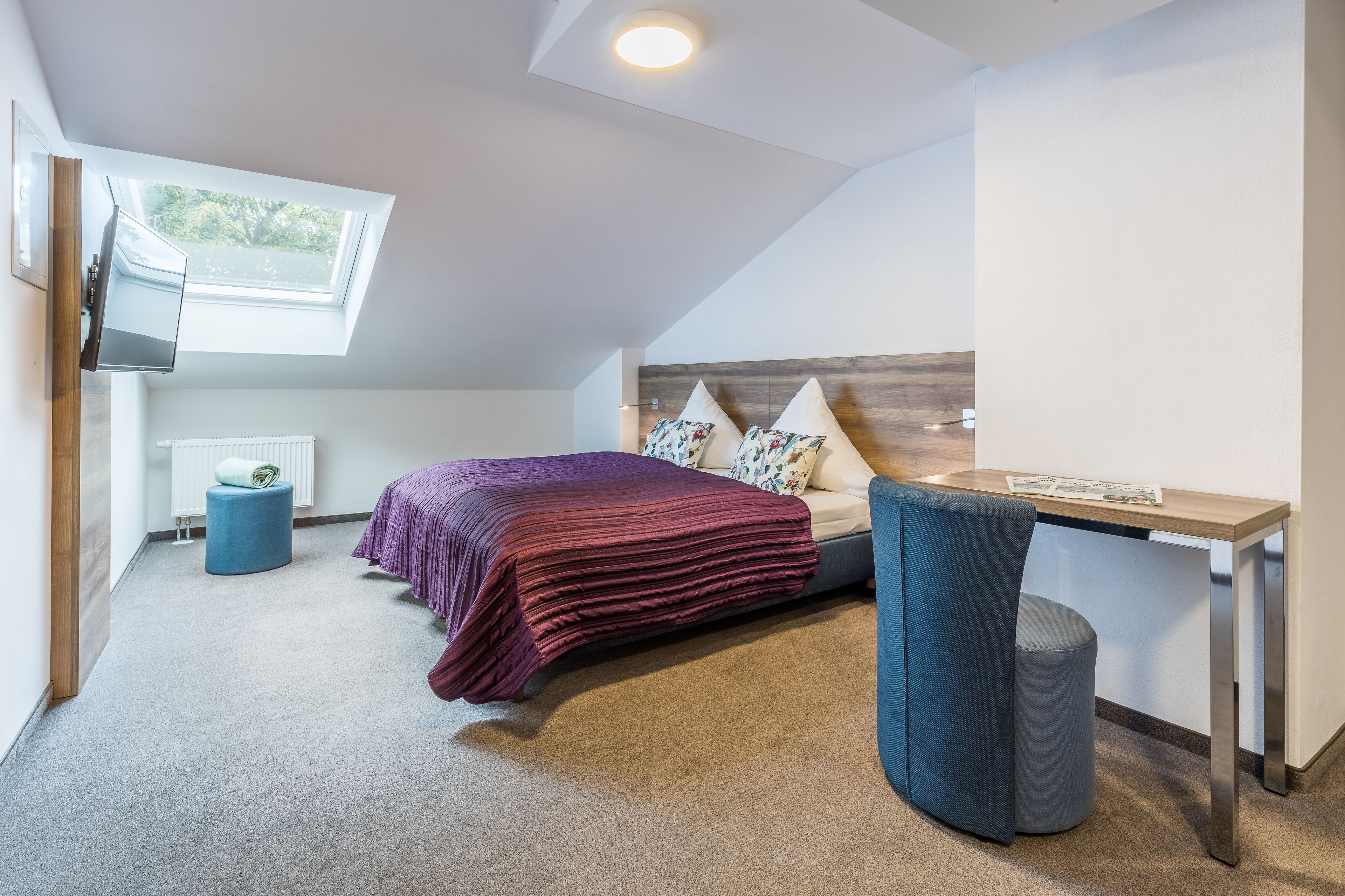 A modern bedroom with a sloped ceiling, a bed with a purple bedspread, a desk, and a chair.