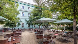 Un café al aire libre con mesas y sillas, rodeado de árboles y un edificio al fondo.