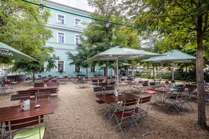 Un café al aire libre con mesas y sillas, rodeado de árboles y un edificio al fondo.