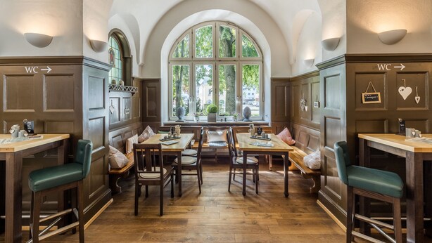 Un comedor con mesas y sillas de madera, un amplio ventanal arqueado y señales indicativas hacia los servicios.