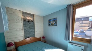 A bedroom with a wooden headboard, a blue bedspread, a wall-mounted light fixture, a small bedside table with a lamp, and a window with a view of parked cars and buildings. - Charming Apartment in Val-Cenis: Ski-In/Ski-Out, Terrace, 4 Guests, Renovated (Lanslevillard)