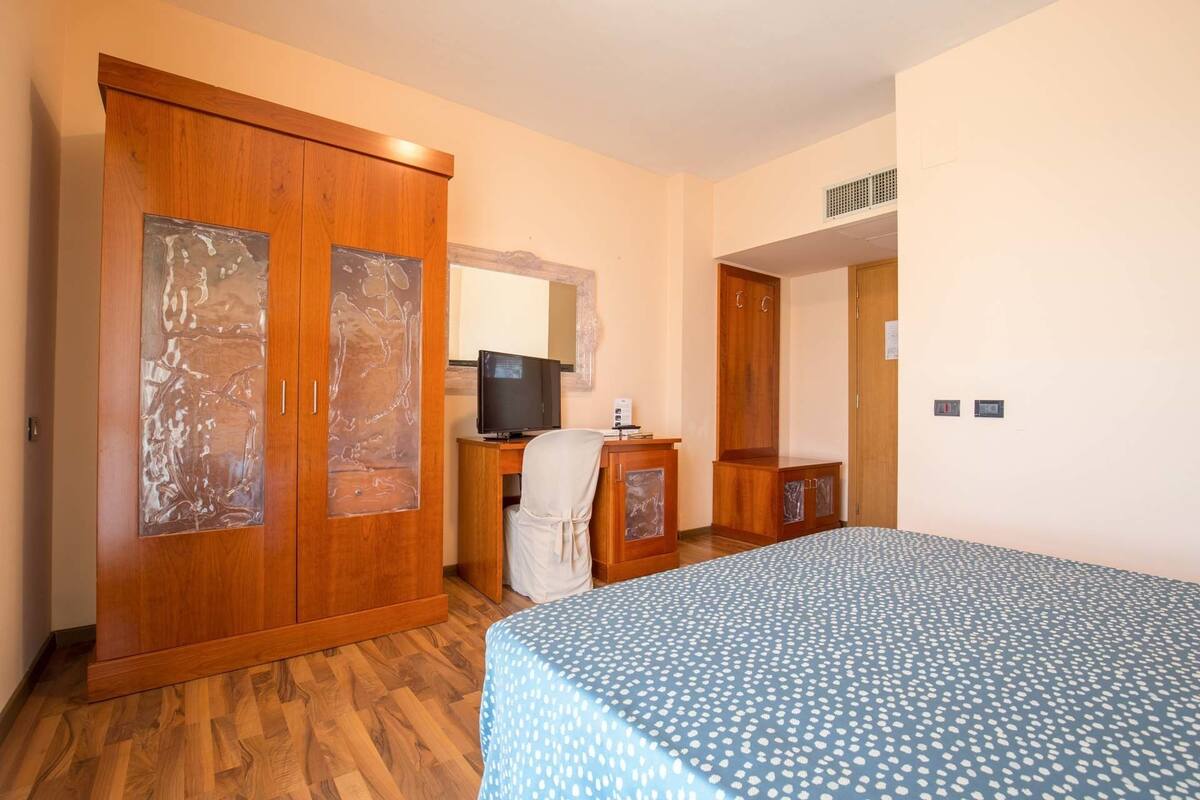 A hotel room with a wooden wardrobe, a desk with a television, a bed with a patterned bedspread, and a mirror on the wall.