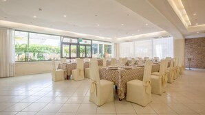 Une salle de banquet spacieuse, avec une longue table dressée pour un événement formel, de grandes fenêtres et un mur décoratif en pierre.