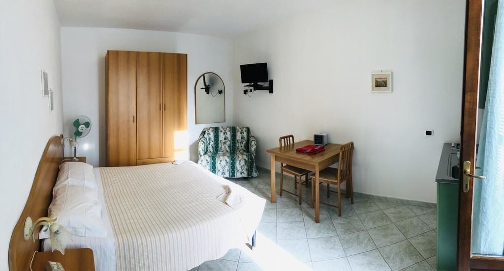 Ein Schlafzimmer mit einem Bett, einem Holzschrank, einem Schreibtisch mit Stuhl, einem an der Wand montierten Fernseher und einem kleinen Tisch mit einem roten Gegenstand.