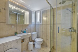 A modern bathroom with a glass-enclosed shower, a toilet, and a washing machine. - Ethrion plus Home experience (Syros)