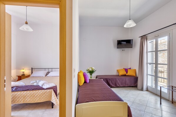 A hotel room with two beds, a TV, a window with curtains, and a bedside table with a lamp. - Ethrion plus Home experience (Syros)