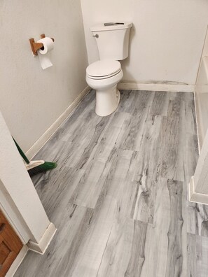 A bathroom with a toilet, a wooden floor, and a broom leaning against the wall.