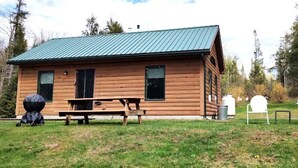 A wooden cabin with a green roof, a picnic table, and two white chairs in the yard.