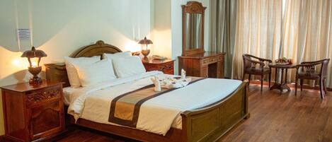 A neatly arranged hotel room with a large bed, bedside tables with lamps, a dresser, a mirror, and a seating area with two chairs and a small table.