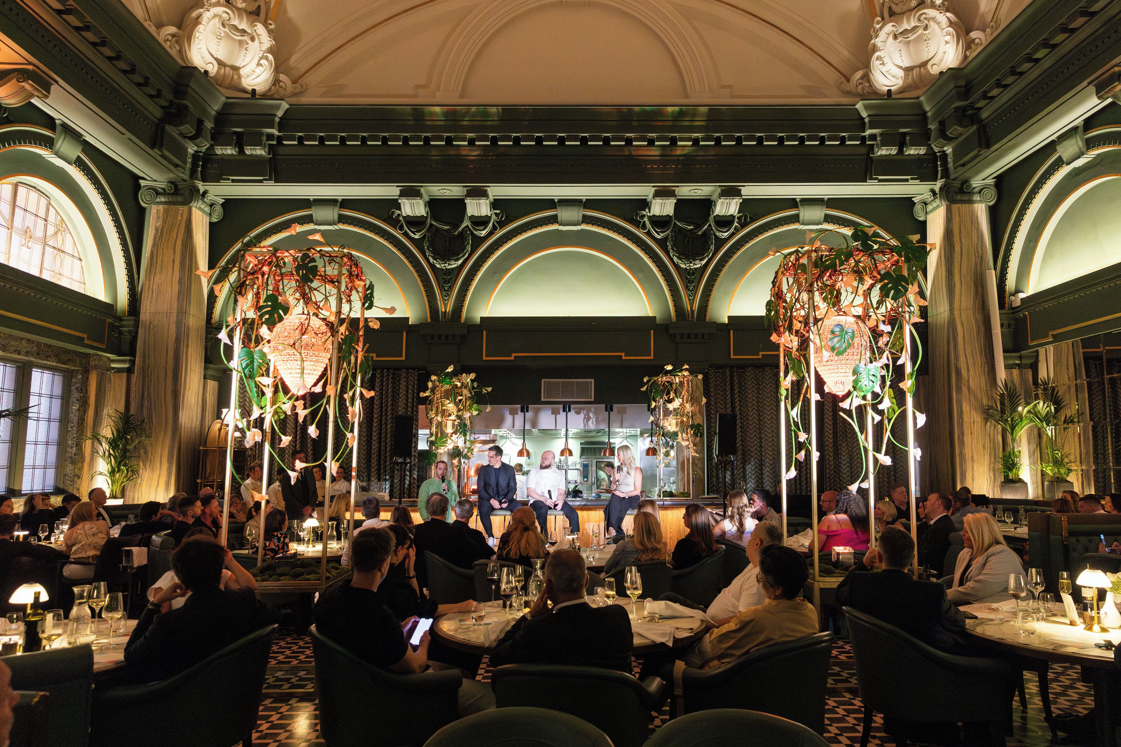 Un comedor formal con mesas dispuestas para invitados, un escenario con público sentado y detalles arquitectónicos ornamentales.