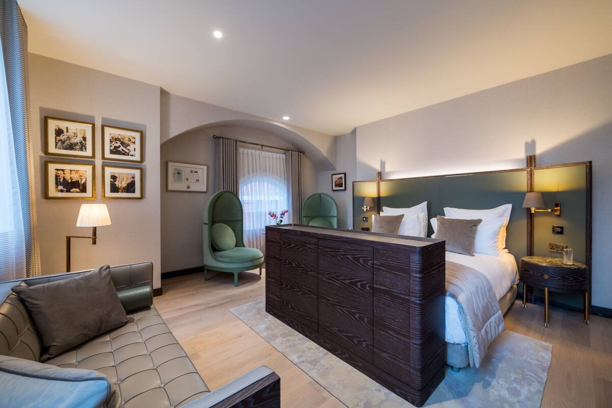 A modern hotel room with a sofa, a green armchair, a bed with a wooden headboard, and a nightstand with a lamp.
