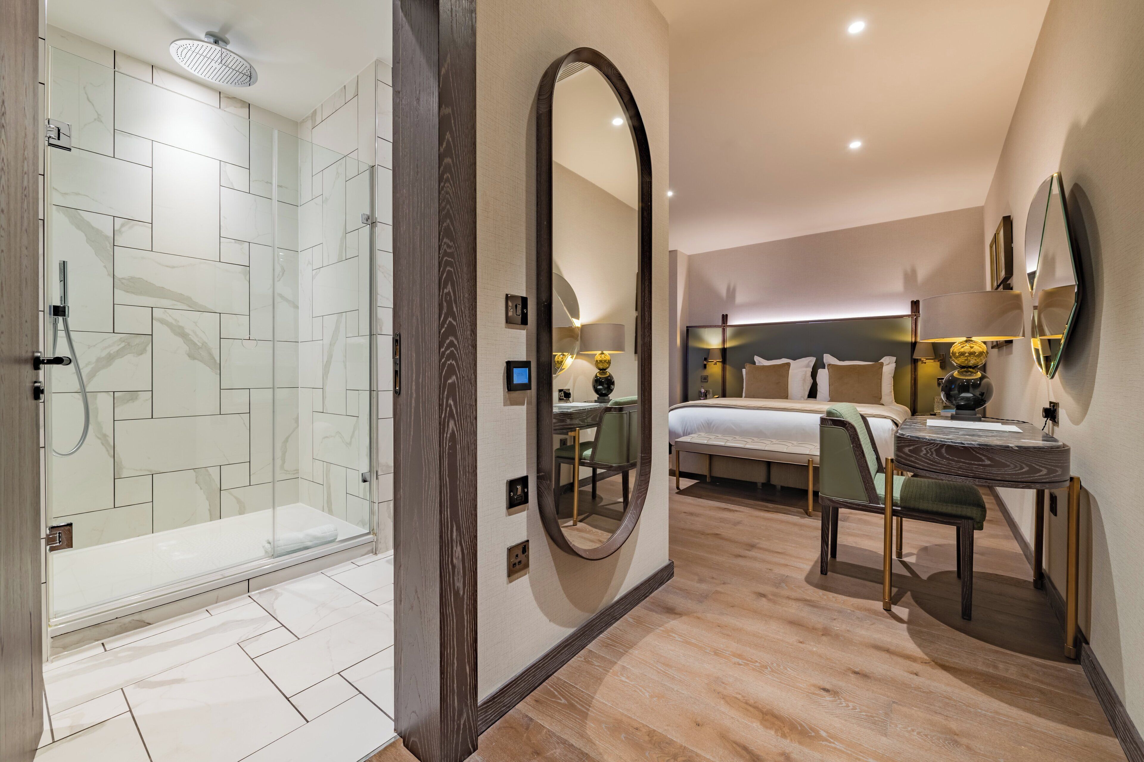Una habitación de hotel moderna con ducha de vidrio, un espejo grande, una cama con cabecera, una mesita de noche con lámpara y una silla.