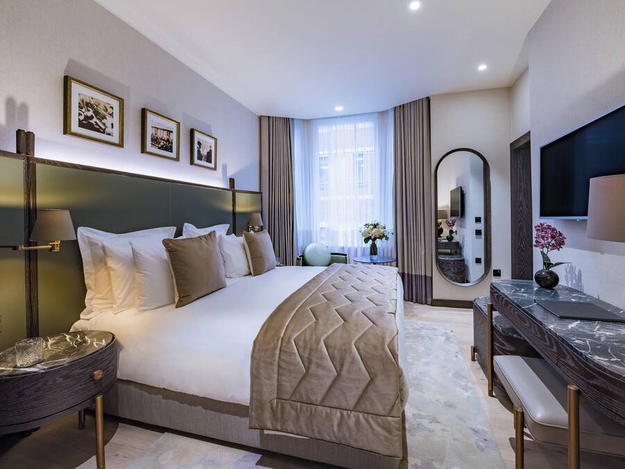 A modern hotel room with a large bed, a desk with a flat-screen TV, a mirror, and framed pictures on the wall.
