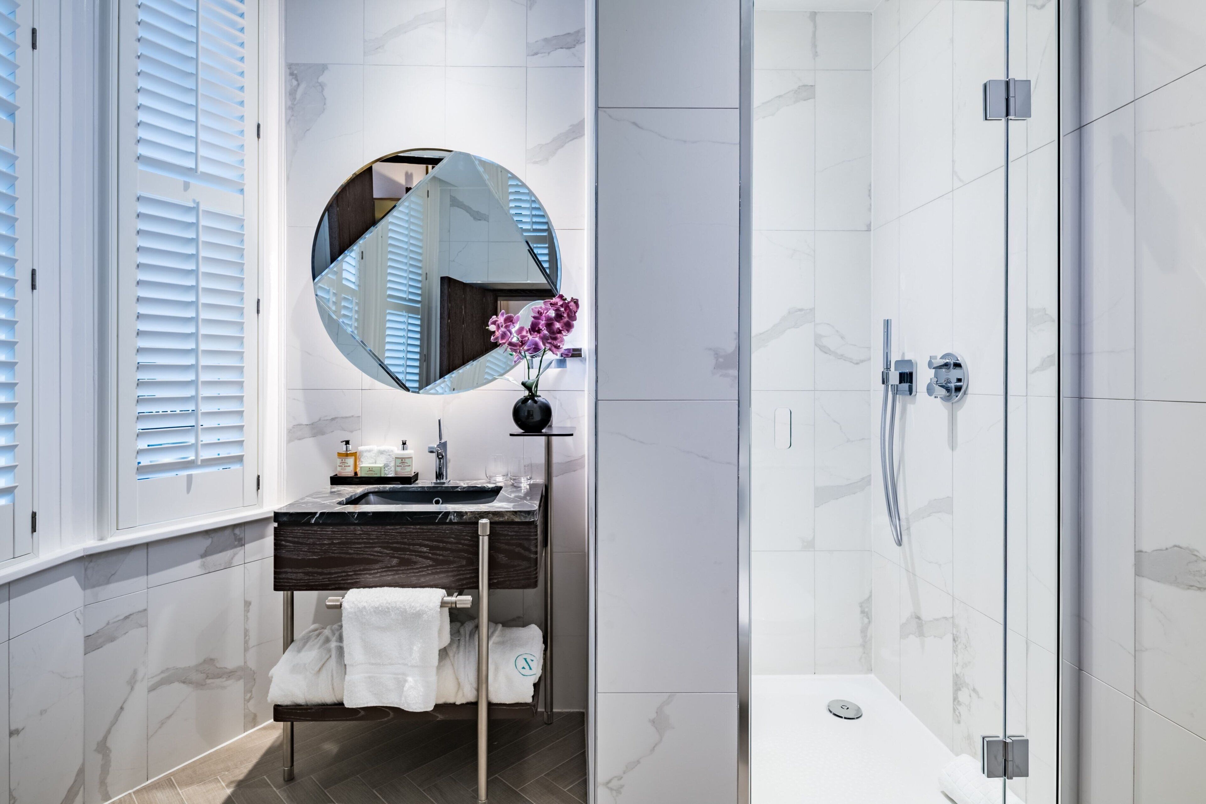 Une salle de bain moderne avec un mur recouvert de carreaux de marbre, un miroir rond, un lavabo doté d’un plan de toilette foncé, une cabine de douche en verre et un porte-serviettes avec des serviettes blanches.