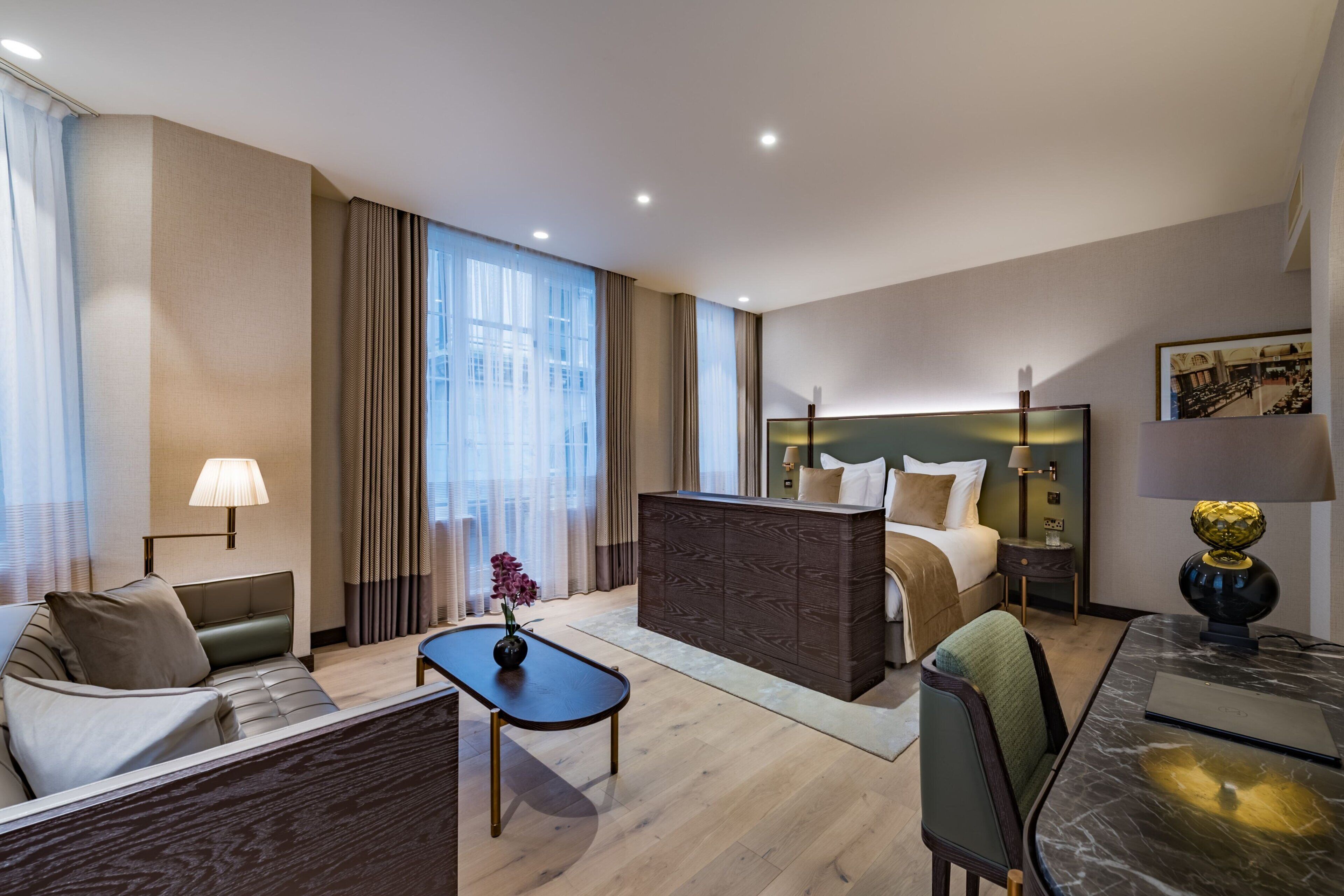 A modern hotel room with a bed, sofa, coffee table, and a desk with a lamp.