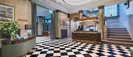 A lobby with a checkered floor, a reception desk, a staircase, and a large chandelier.