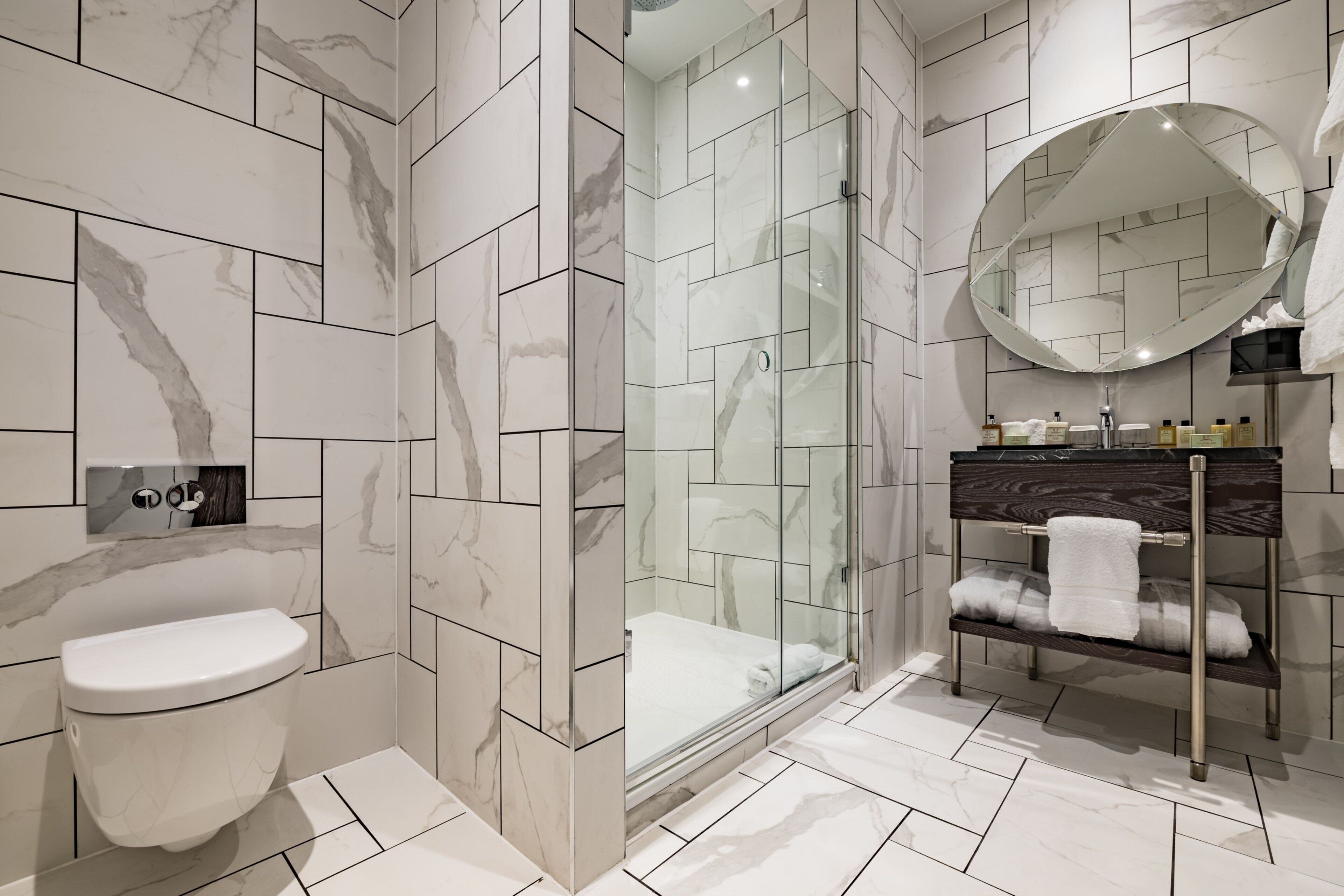 Un baño moderno con cabina de ducha de vidrio, pared con azulejos de mármol, inodoro blanco y tocador con espejo y toallero.