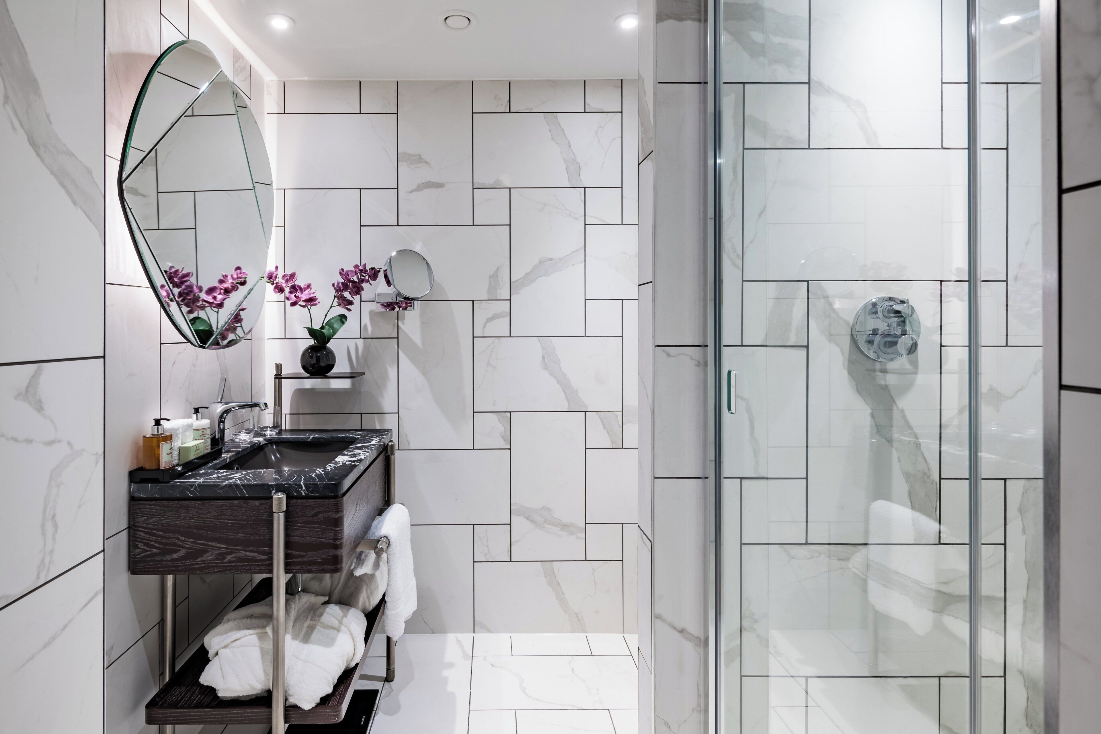 A modern bathroom with a glass-enclosed shower, a marble-tiled wall, a vanity with a dark countertop, and a large mirror.
