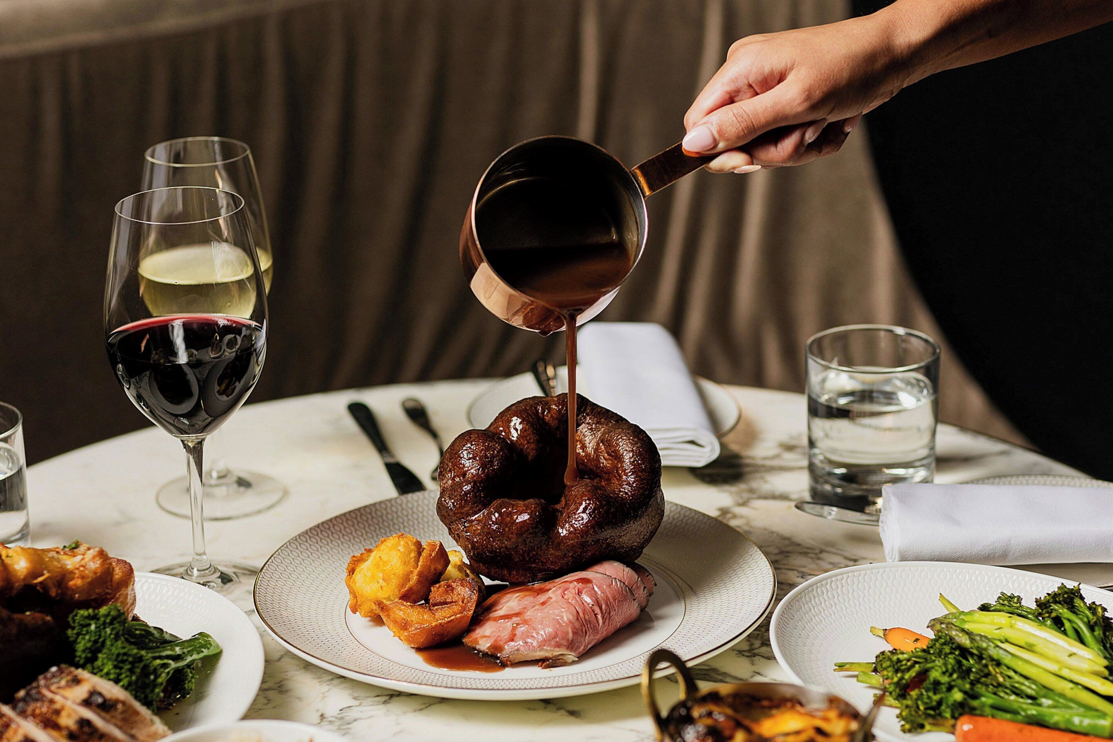 A dining table with plates of food, a glass of red wine, and a person pouring a dark liquid into a bowl.